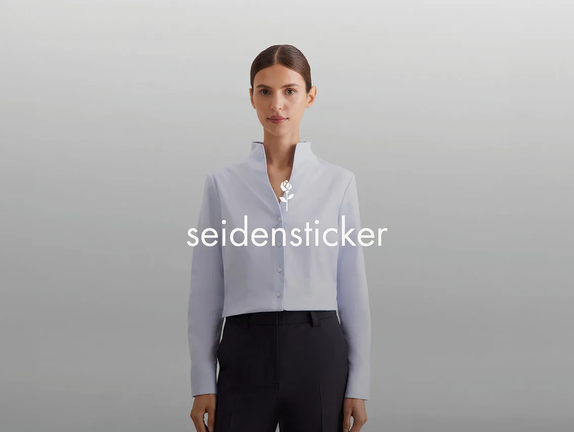 Žena s vlasy staženými dozadu má na sobě světle modrou zkrácenou košili s vysokým límcem a tmavé kalhoty. Na hrudi je bílé logo „seidensticker“ s růží.